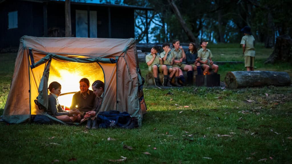 Tudor House students sitting in a tent playing cards, other students sitting on a log and toasting marshmallows