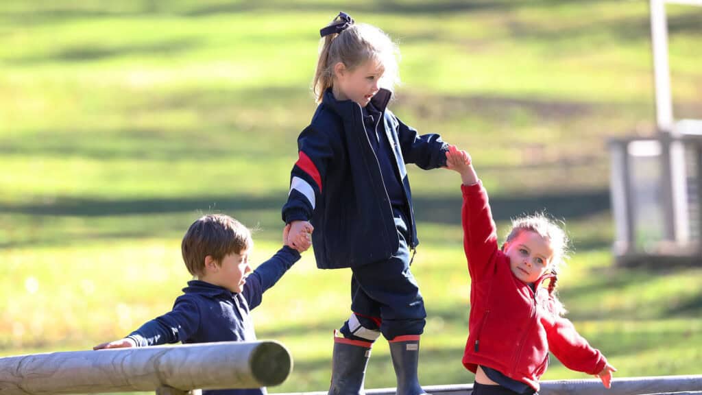 Tudor House pre-kindergarten student walking across a beam in gumboots whole two other students holde her hands